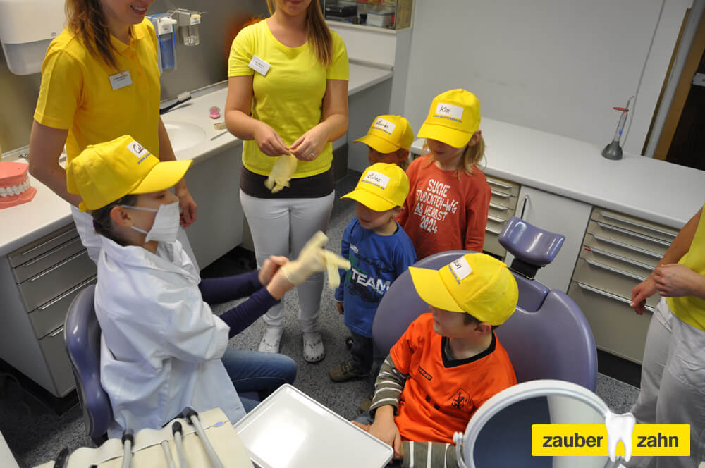 Kinder mit gelben Kappen stehen um einen zahnärztlichen Behandlungsstuhl.