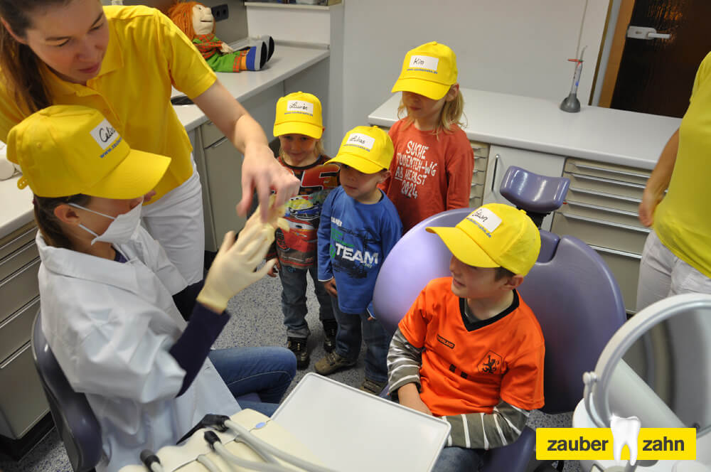 Kinder mit gelben Kappen stehen um einen zahnärztlichen Behandlungsstuhl.