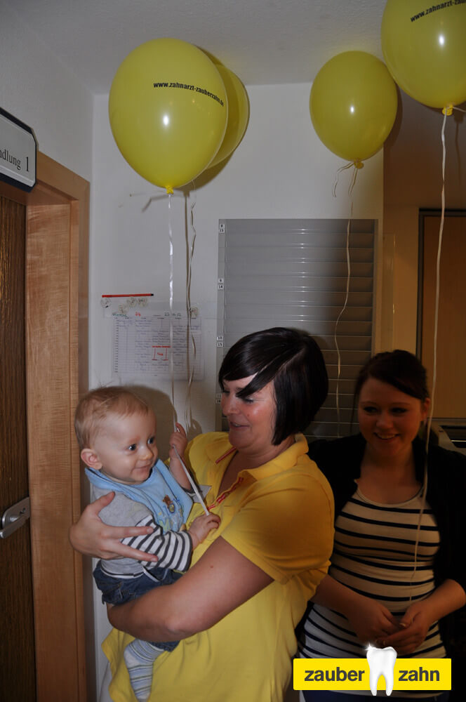 Mitarbeiterin hält ein Kleinkind unter gelben Luftballons auf dem Arm.