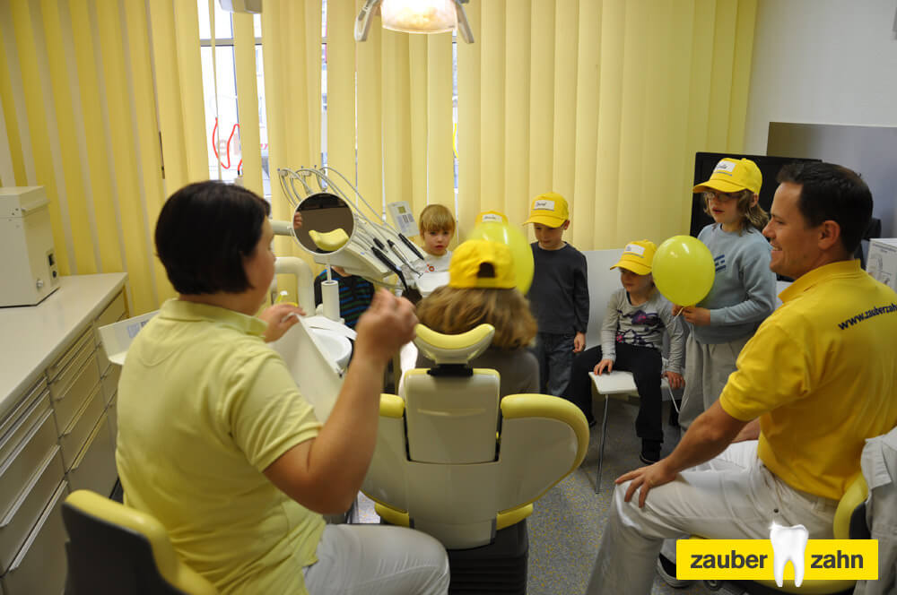 Mitarbeiter spricht mit einer Gruppe von Kindern mit gelben Kappen im Behandlungszimmer.