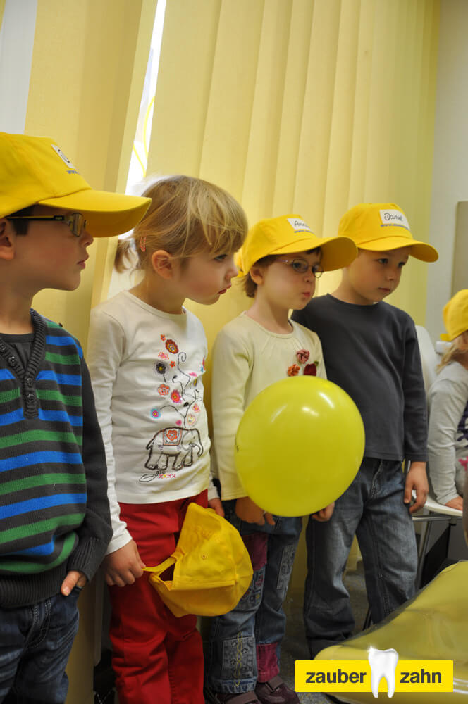 Mehrere Kinder mit gelben Kappen stehen gemeinsam neben einem gelben Luftballon.