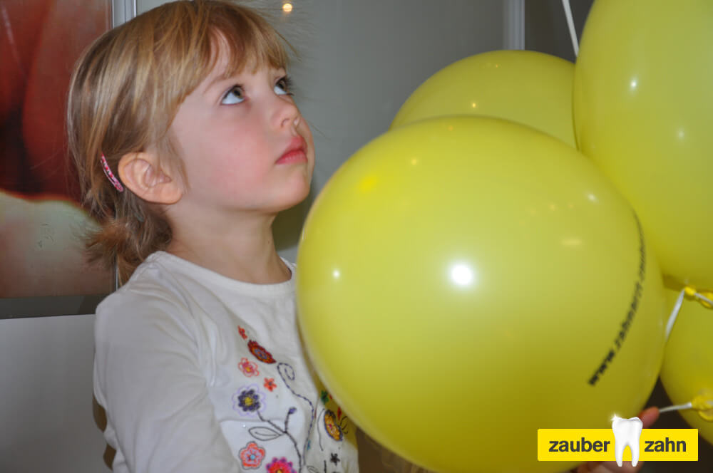 Kind steht neben mehreren gelben Luftballons und blickt nach oben.