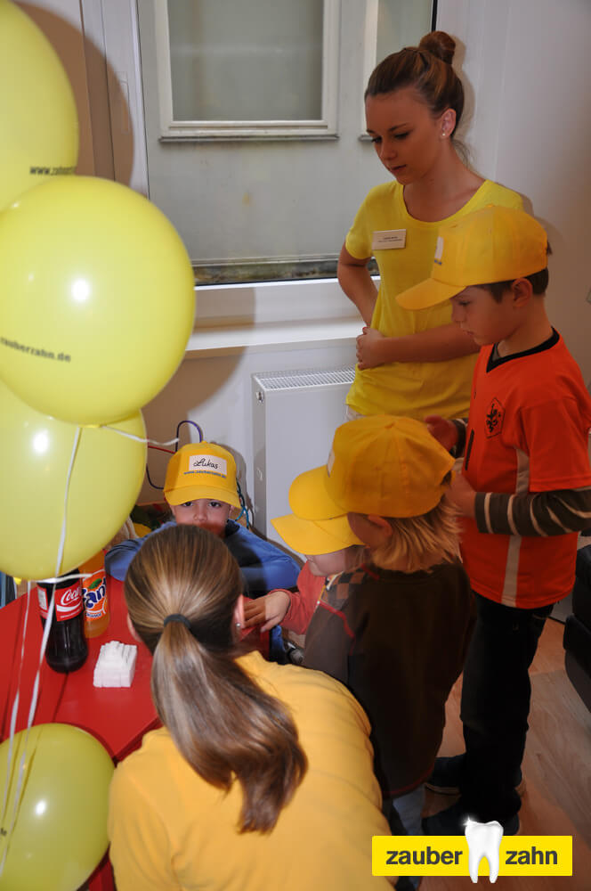 Gruppe von Kindern mit gelben Kappen und gelben Luftballons im Behandlungszimmer.