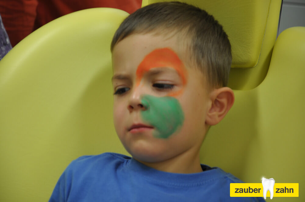 Kind mit orange-grüner Gesichtsbemalung sitzt auf einem Behandlungsstuhl.