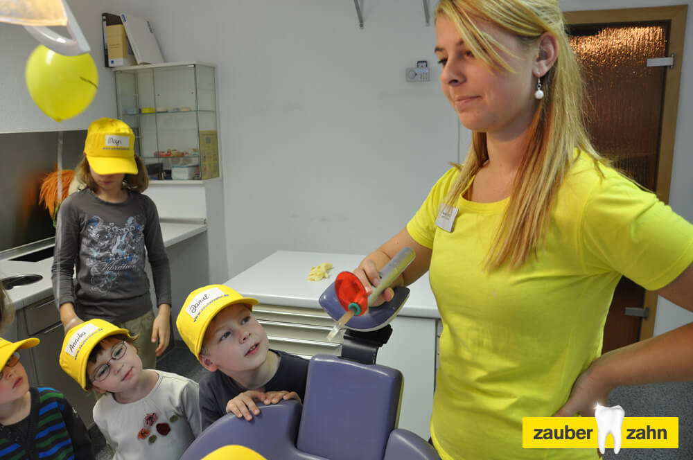 Mitarbeiterin zeigt Kindern ein zahnärztliches Instrument.