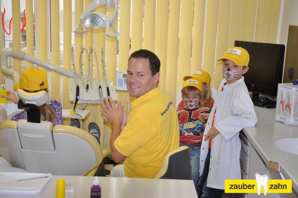 Mitarbeiter sitzt an der zahnärztlichen Behandlungseinheit, während Kinder mit gelben Kappen danebenstehen.