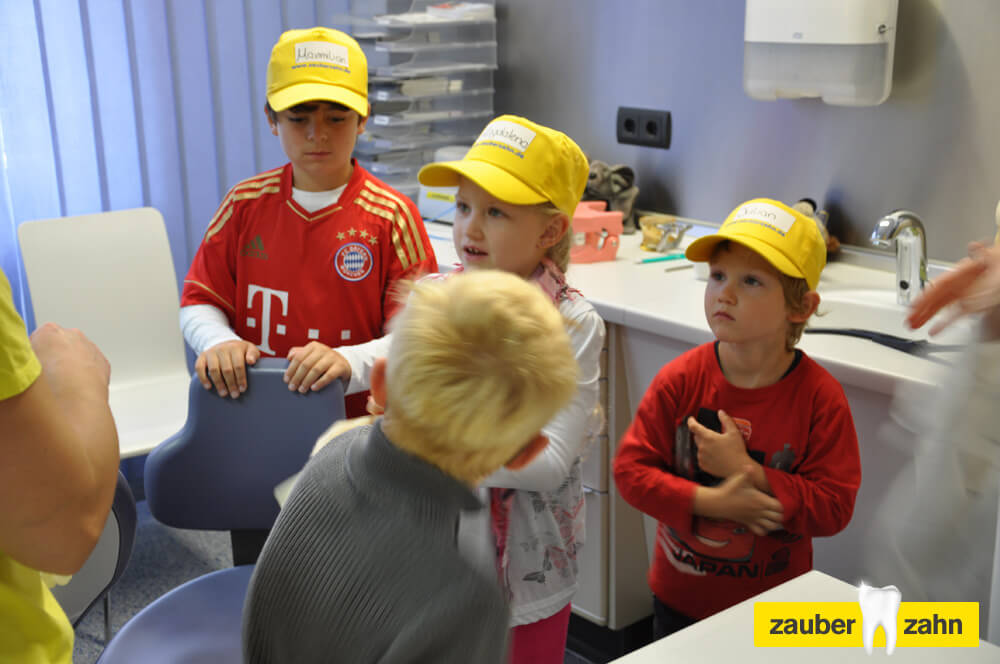 Mehrere Kinder mit gelben Kappen stehen gemeinsam im Behandlungszimmer.