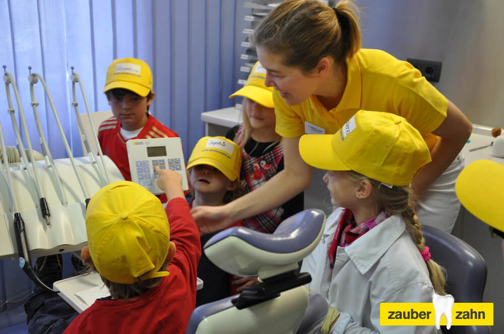 Mitarbeiterin zeigt Kindern mit gelben Kappen ein zahnärztliches Instrument.