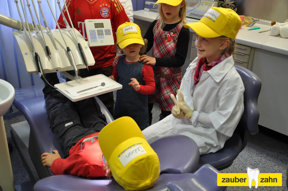 Kinder mit gelben Kappen stehen um ein Kind auf dem zahnärztlichen Behandlungsstuhl.