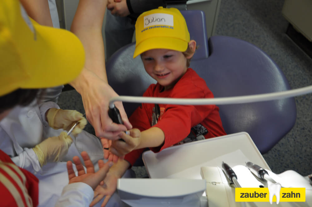 Kind mit gelber Kappe probiert ein zahnärztliches Instrument an einer Hand aus.