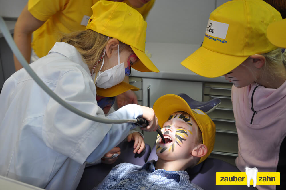 Kind mit bemaltem Gesicht liegt auf einem Behandlungsstuhl, während ein anderes Kind ein zahnärztliches Instrument hält.