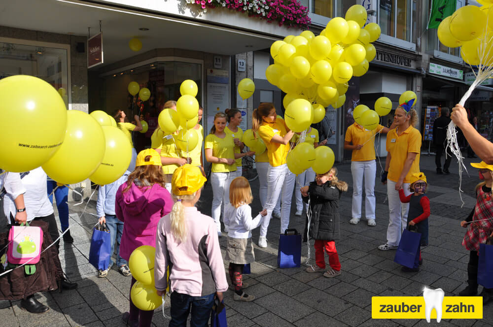 Kinder und Mitarbeiterinnen mit gelben Luftballons vor einem Geschäft.