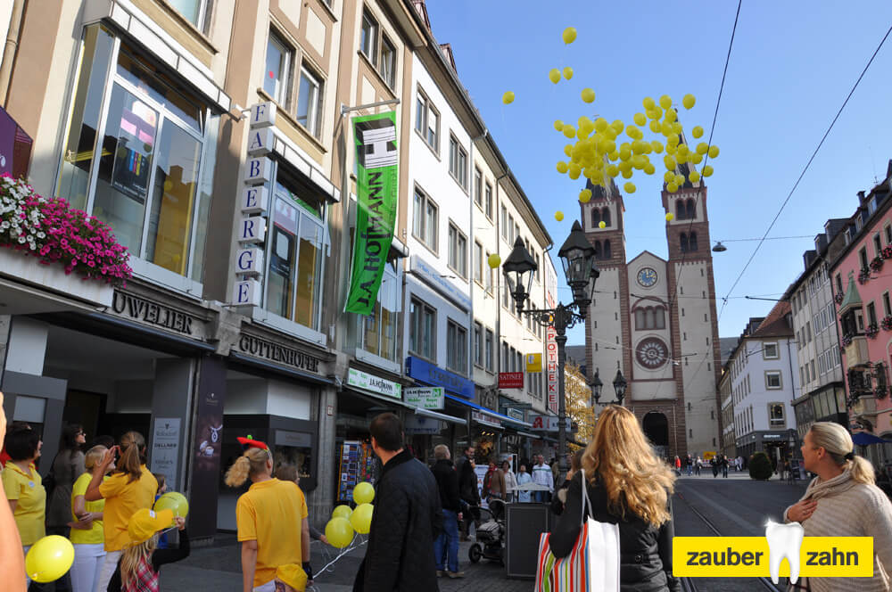Gelbe Luftballons steigen über einer Fußgängerzone mit Kirchtürmen in den Himmel.