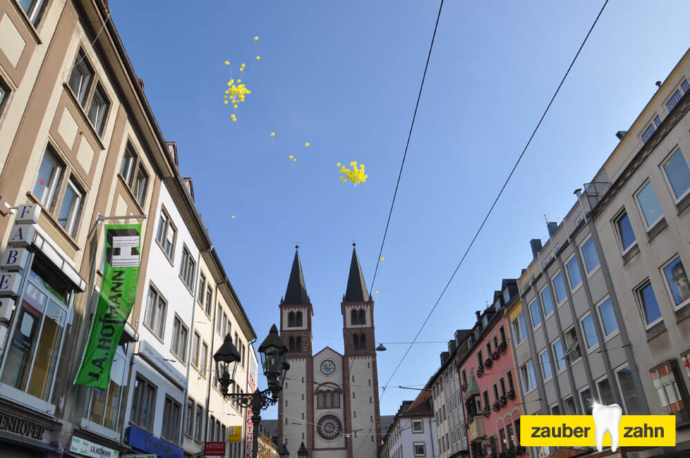 Gelbe Luftballons steigen über einer Straße mit zwei Kirchtürmen in den Himmel.