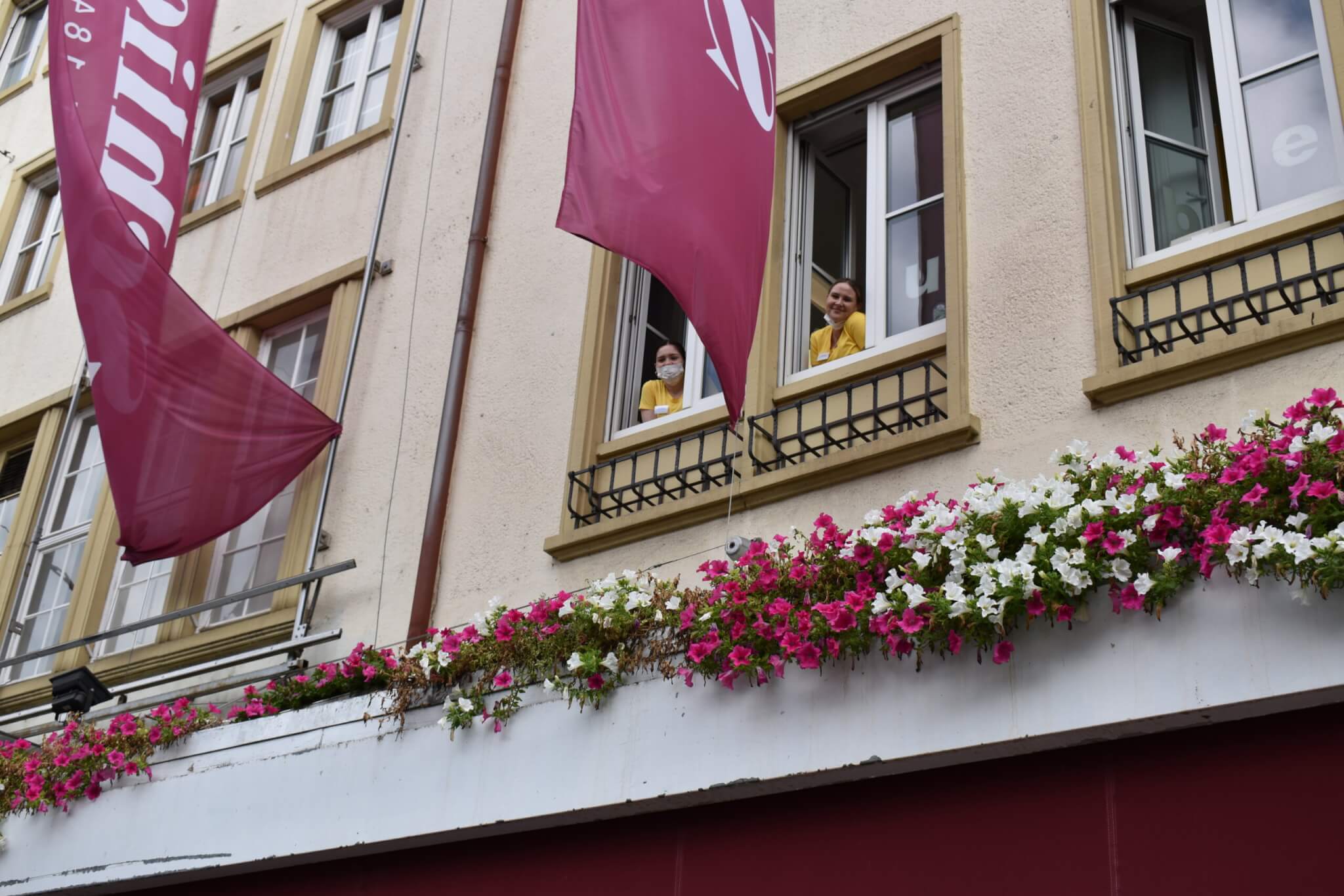 Zwei Mitarbeiterinnen der Zahnarztpraxis schauen aus einem geöffneten Fenster über einer mit Blumen geschmückten Fassade.