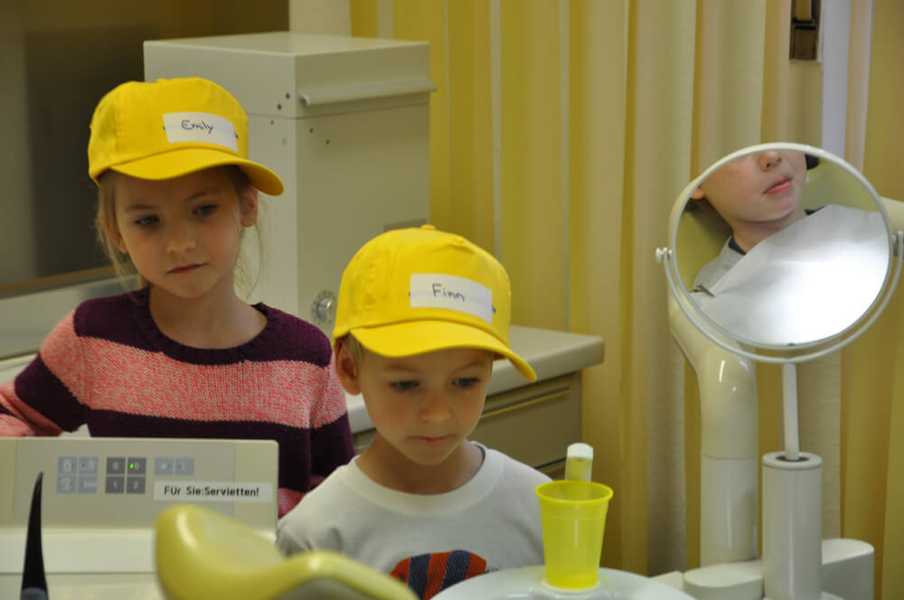Zwei Kinder mit gelben Kappen stehen an einer zahnärztlichen Behandlungseinheit.