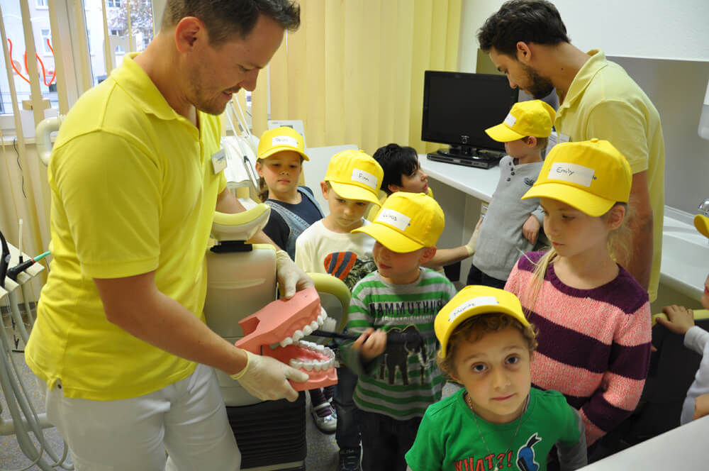 Mitarbeiter zeigt Kindern mit gelben Kappen ein Gebissmodell.