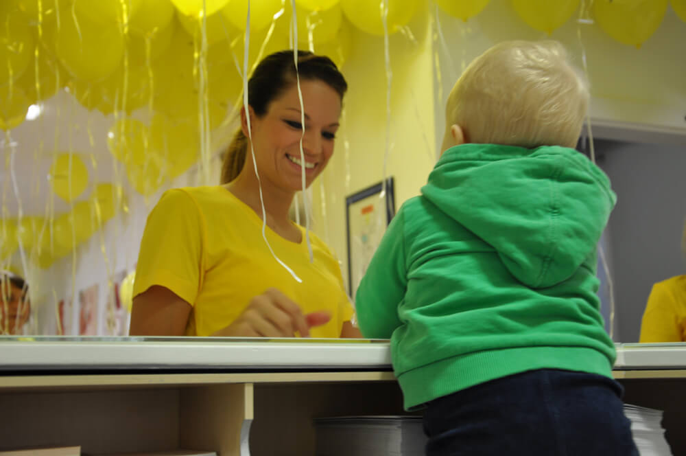 Mitarbeiterin steht einem Kleinkind im mit gelben Luftballons geschmückten Empfangsbereich gegenüber.