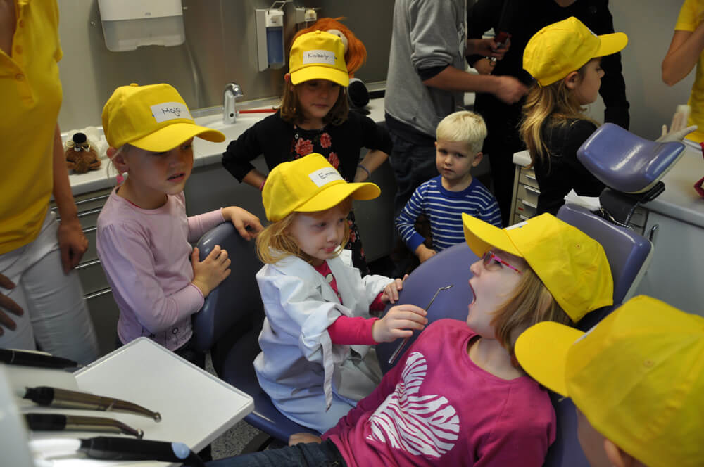 Kinder mit gelben Kappen beobachten ein Kind auf dem Behandlungsstuhl.