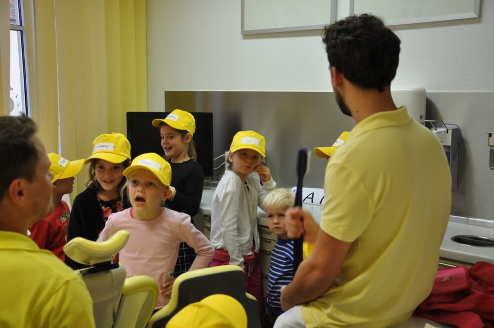 Mitarbeiter spricht mit mehreren Kindern mit gelben Kappen im Behandlungszimmer.