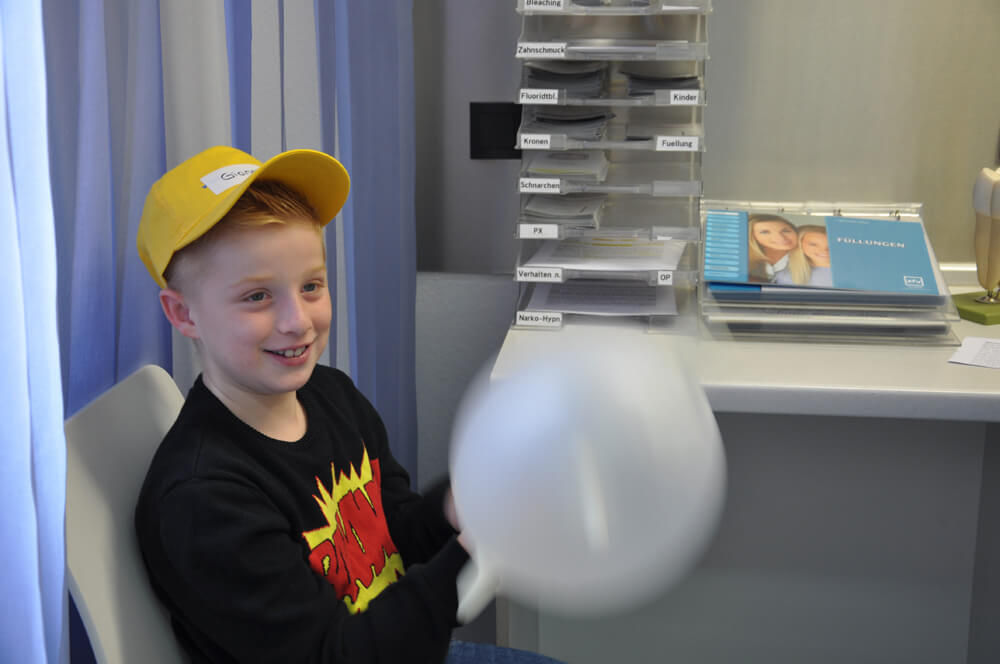 Kind mit gelber Kappe hält einem weißen Luftballon im Behandlungszimmer.