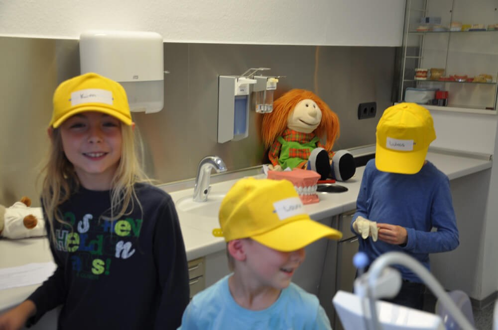 Kinder mit gelben Kappen im Behandlungszimmer neben einer Handpuppe.