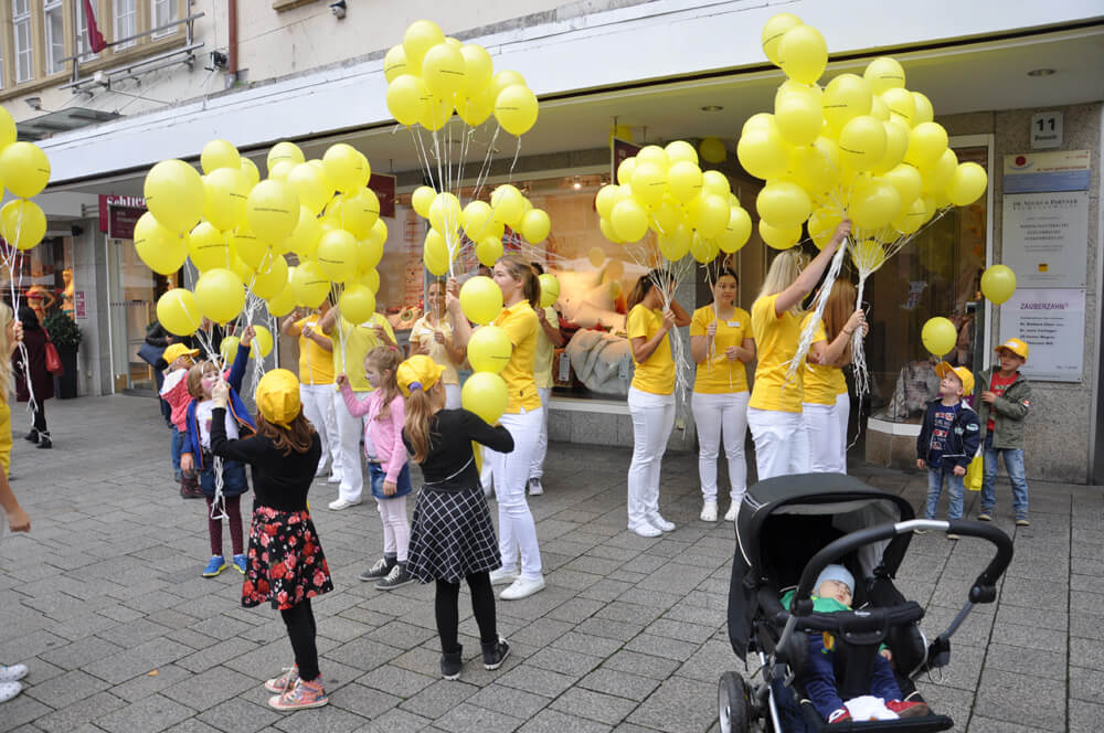 Kinder und Erwachsene halten zahlreiche gelbe Luftballons vor einem Gebäude.