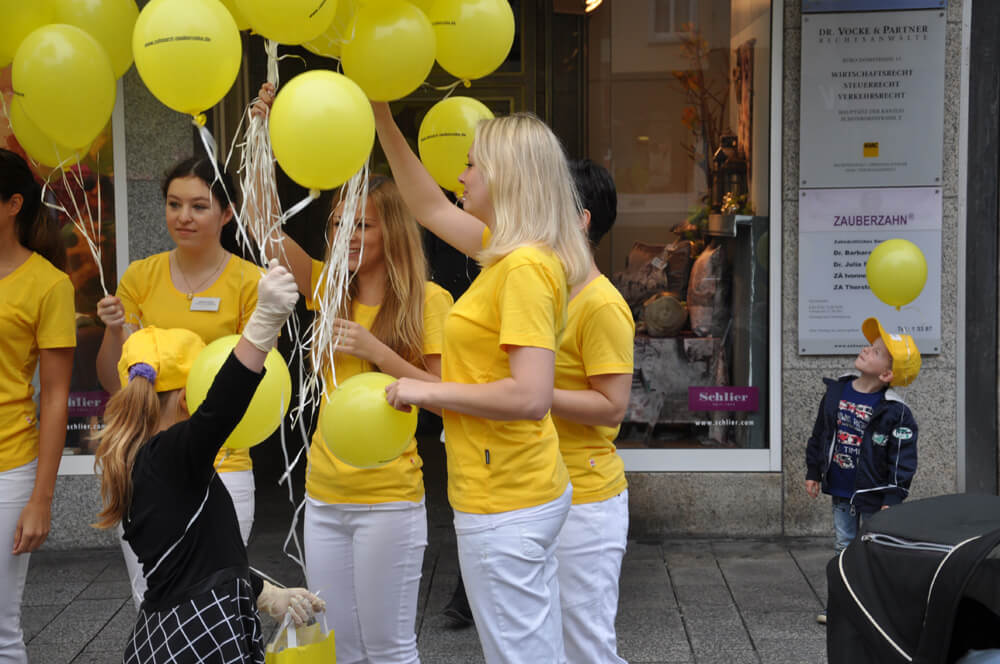 Mitarbeiterinnen halten gelbe Luftballons vor einem Gebäude.