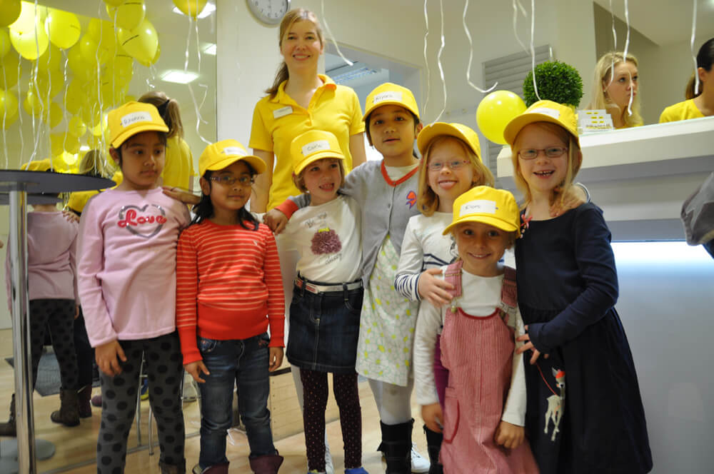Gruppe von Kindern mit gelben Kappen steht gemeinsam im Behandlungszimmer und machen ein Foto.