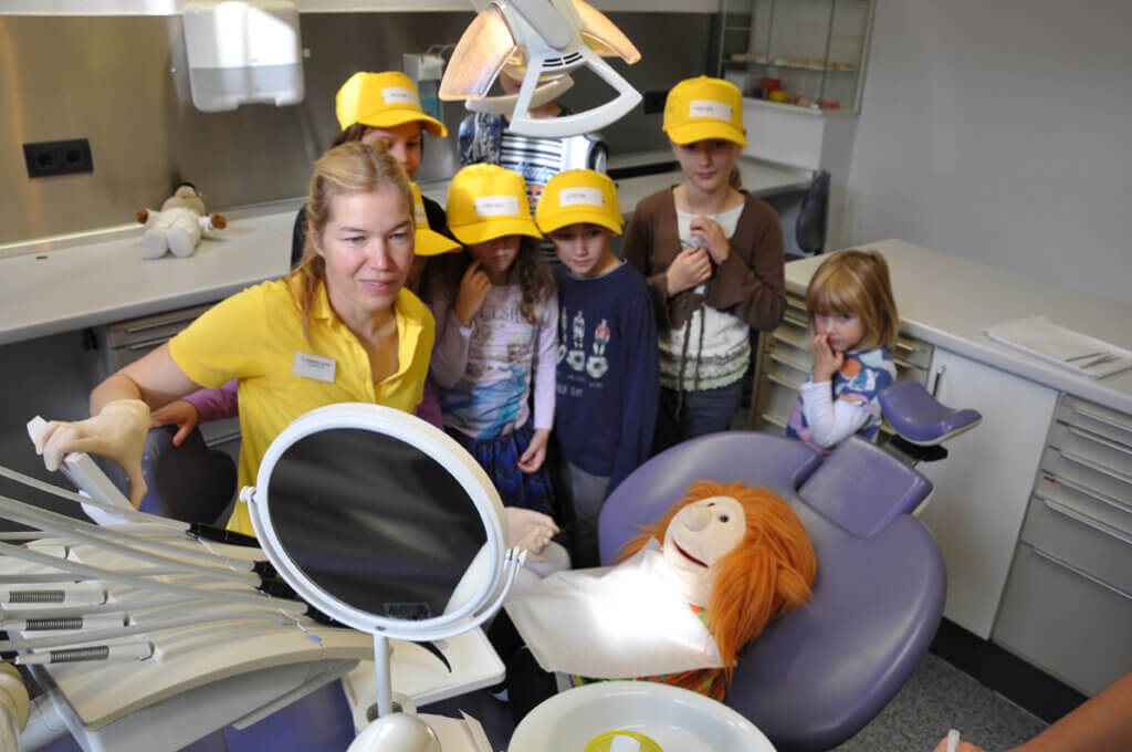 Mitarbeiterin zeigt Kindern mit einer Handpuppe eine zahnärztliche Behandlung.