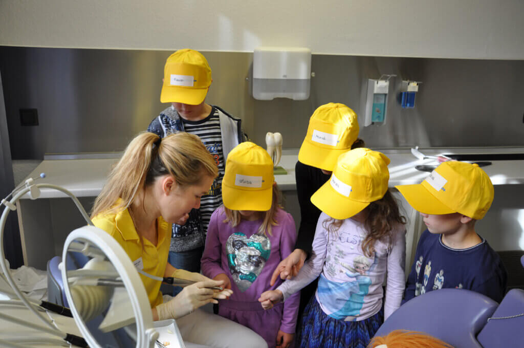 Mitarbeiterin zeigt Kindern ein zahnärztliches Instrument.