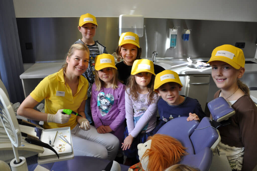 Mitarbeiterin und Kinder mit gelben Kappen im Behandlungszimmer und lächeln.