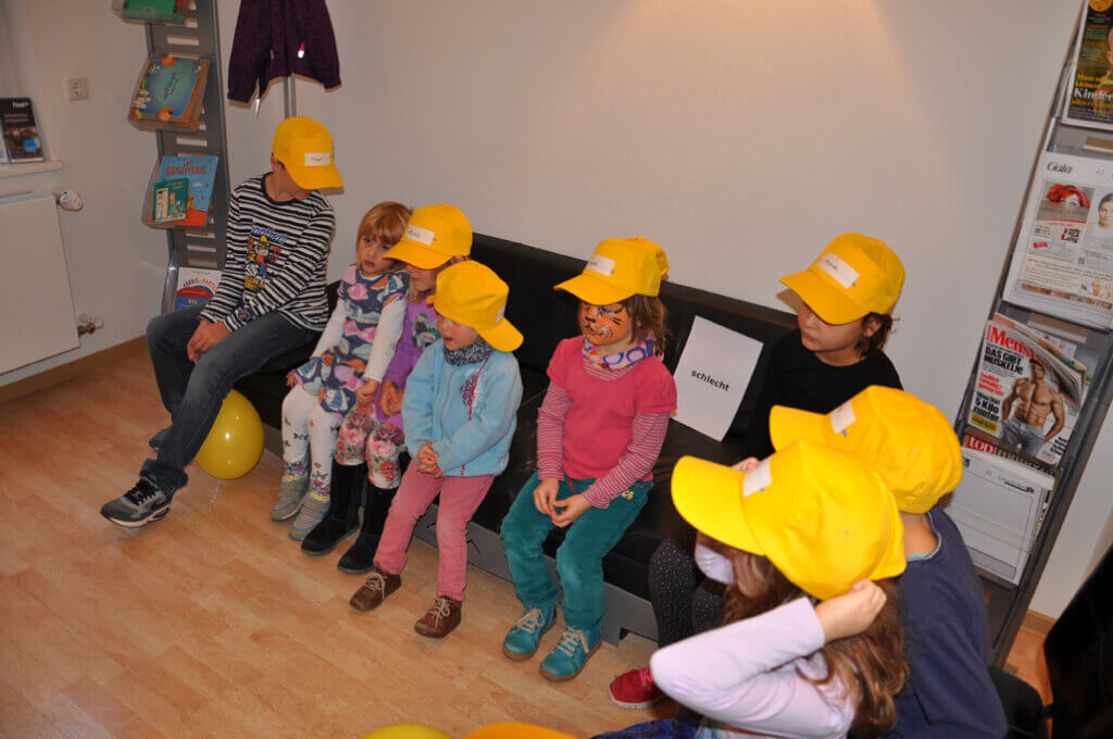Kinder mit gelben Kappen sitzen gemeinsam im Wartebereich.