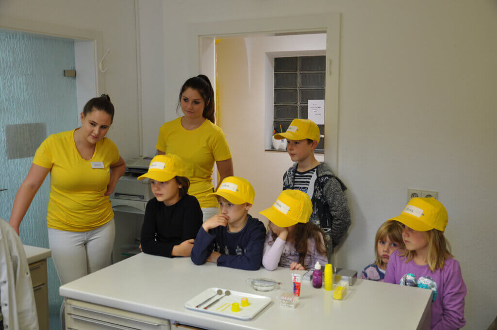 Kinder sitzen gemeinsam mit zwei Mitarbeiterinnen im Behandlungszimmer.